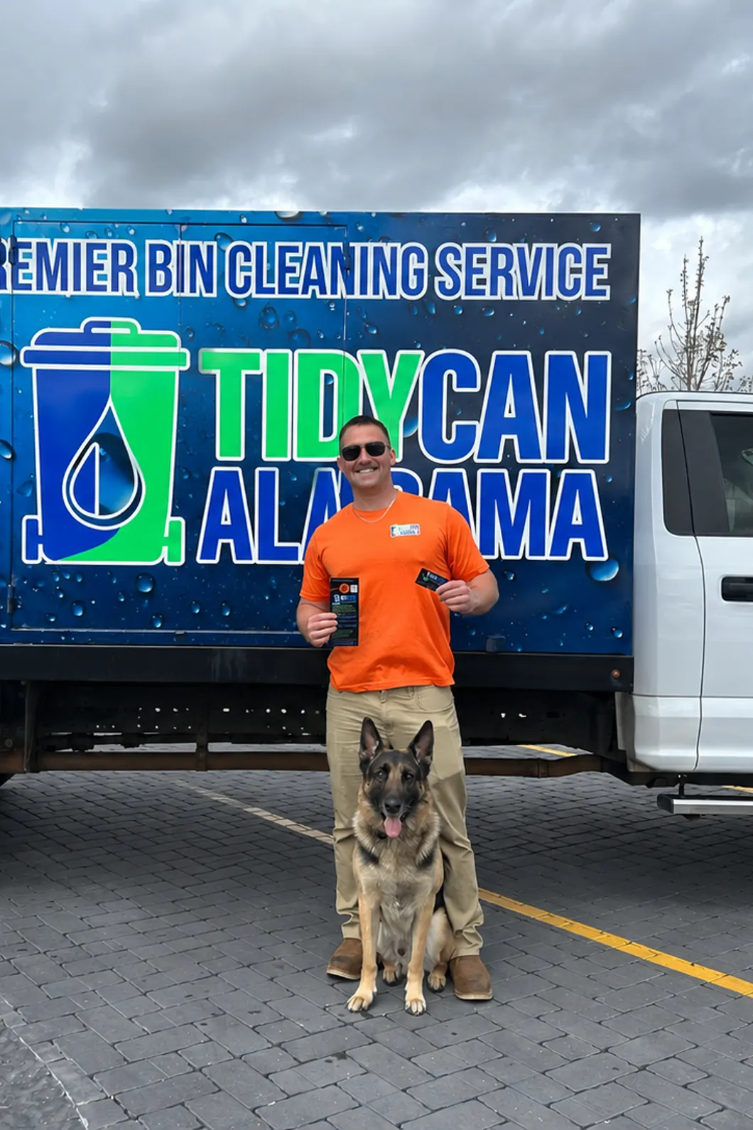 Jason Hogan, owner and operator of TidyCan Alabama, standing with a German shepherd in front of the TidyCan bin cleaning truck